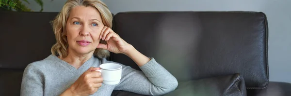 Mulher idosa sentada no sofá em sua sala de estar e segurando uma xícara de café . — Fotografia de Stock