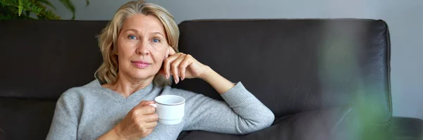 Mulher idosa sentada no sofá em sua sala de estar e segurando uma xícara de café . — Fotografia de Stock