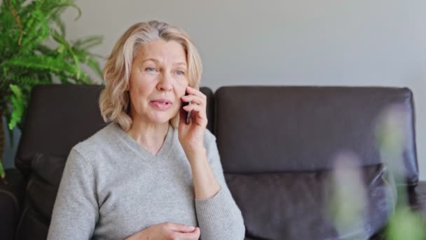 Gammal kvinna som använder mobiltelefon när hon sitter på soffan hemma. — Stockvideo