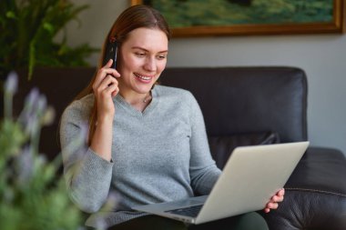 Kanepede dizüstü bilgisayarıyla oturan ve evde telefonla konuşan mutlu kadın..
