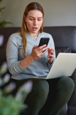 Kanepede dizüstü bilgisayarıyla oturan ve evde telefonla konuşan mutlu kadın..