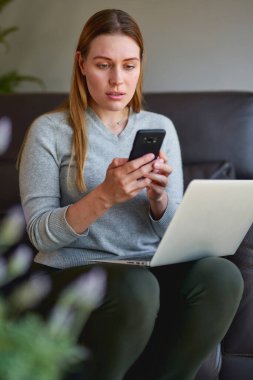 Kanepede dizüstü bilgisayarıyla oturan ve evde telefonla konuşan mutlu kadın..