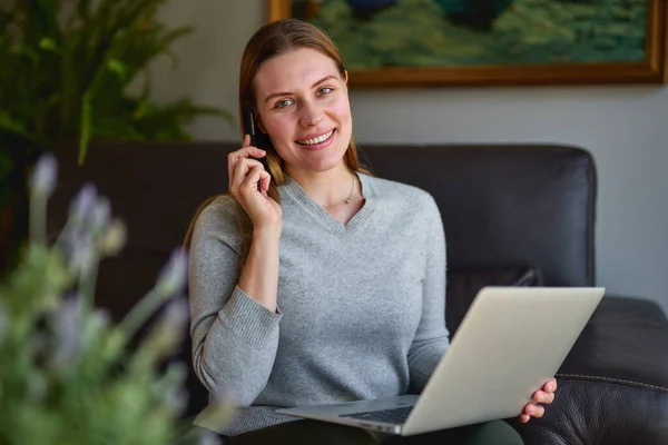 소파에 노트북을 놓고 앉아서 집에서 전화 통화를 하는 행복 한 여성. — 스톡 사진