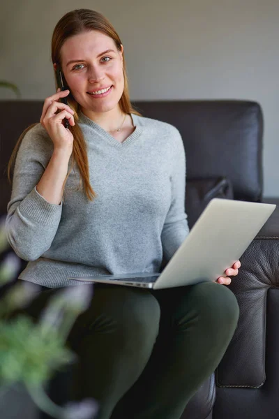 Ευτυχισμένη γυναίκα κάθεται στον καναπέ με φορητό υπολογιστή και μιλάμε στο τηλέφωνο στο σπίτι. — Φωτογραφία Αρχείου