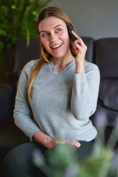 Happy woman sitting on sofa with laptop and talking on phone at home. — Stock Photo, Image