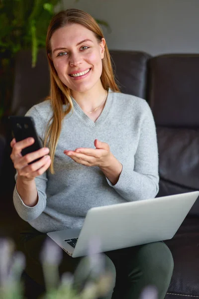 Mulher feliz sentada no sofá com laptop e falando ao telefone em casa . — Fotografia de Stock