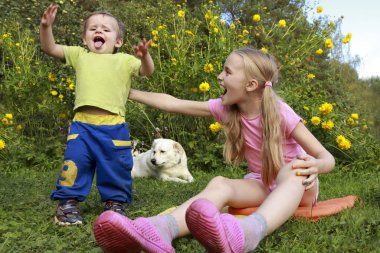 Happiness of life.A beautiful girl plays and has fun with a baby boy, clap their hands and laugh loudly, a mongrel pet lies next to the background of green plants and blue sky in summer. clipart