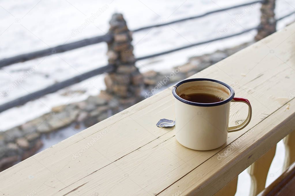 Enamel mug with strong tea and tea bag on a wooden fence on a snowy natural background.