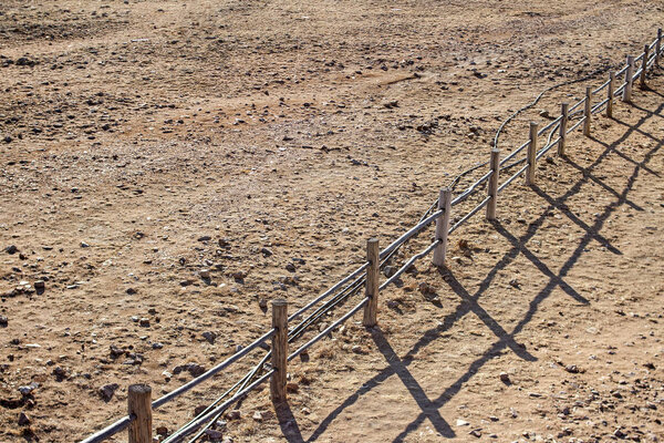 Authentic wooden fence in the countryside casts a shadow on the sandy soil during the sunrise.