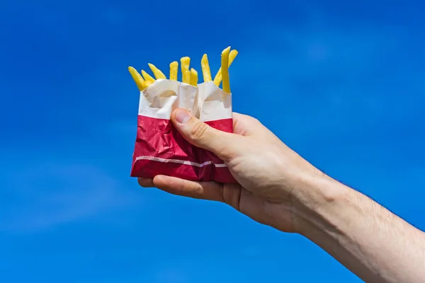 Crispy french fries in a paper bag in a male hand on a blue sky background. — Stock Photo, Image