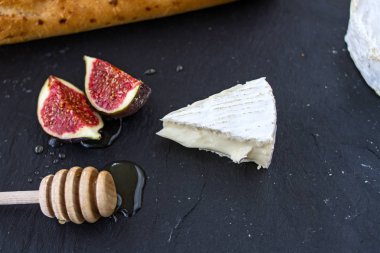 Camembert peynirinin üçgen iştah açıcı parçası, bal içinde incir parçaları ve grafit arka planda bal ve baget için ahşap kaşık.