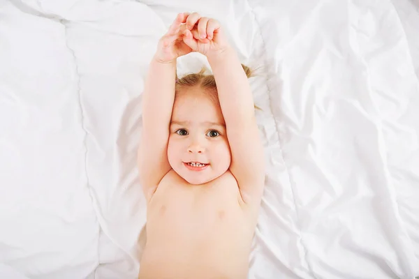 Μικρό παιδί ξαπλωμένος στο κρεβάτι στο σπίτι και το τέντωμα. 2 έτη παλιά μωρό στηρίζεται σε άσπρη κουβέρτα. — Φωτογραφία Αρχείου