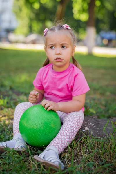 Χαριτωμένο παιδί κάθεται στο γρασίδι στο πάρκο, παίζοντας με την πράσινη μπάλα και χαμογελαστό κοριτσάκι — Φωτογραφία Αρχείου