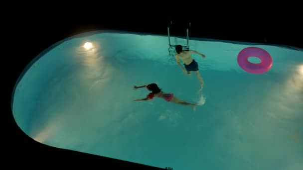 Rapaz e rapariga banham-se na piscina à noite. Crianças diversão nadar na água crianças — Vídeo de Stock