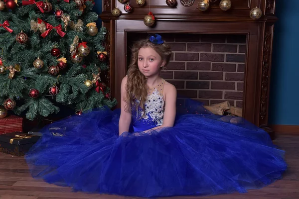 Girl in blue dress sits by fireplace at christmas time — Stock Photo, Image