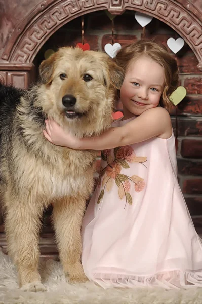Chica en un vestido rosa con un perro mestizo junto a la chimenea — Foto de Stock