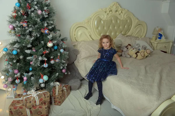 파란 드레스를 입고 반짝 이는 아이가 크리스마스 옆 침대에 앉아 있다 — 스톡 사진