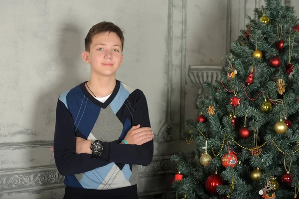 Teenager boy in a sweater stands next to a Christmas tree — Stock Photo, Image