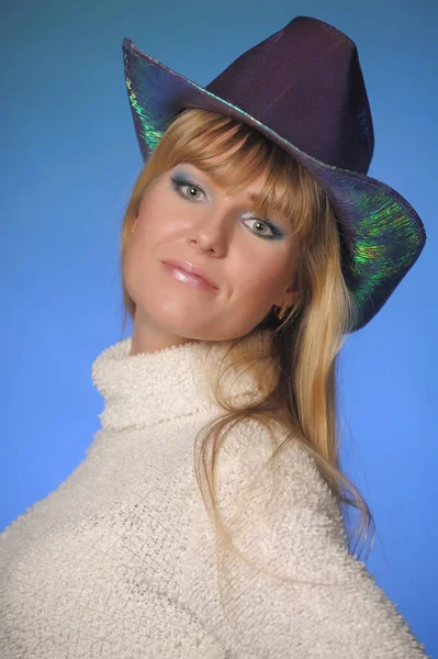 Dlouhosrstá blondýna v ateliéru na modrém pozadí v klobouku — Stock fotografie