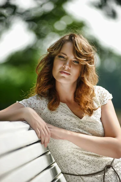 Young brunette in summer in a white dress sits on a park bench — Stock Photo, Image