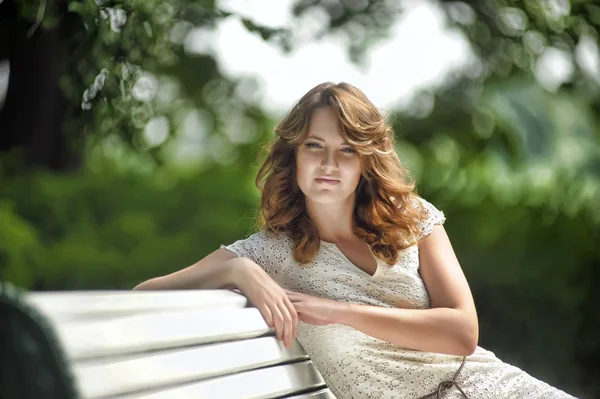Jovem morena no verão em um vestido branco senta-se em um banco de parque — Fotografia de Stock