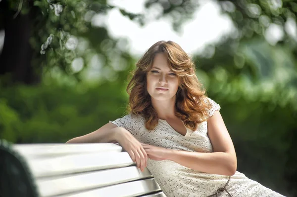 Young brunette in summer in a white dress sits on a park bench — 스톡 사진