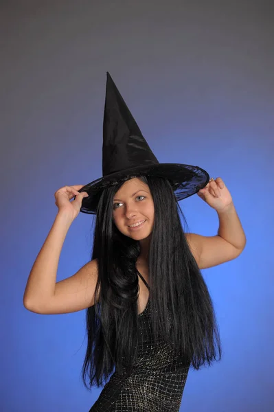 Brunette girl in a black dress and a witch hat on a blue backgro — Stock Photo, Image