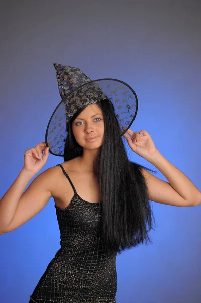 Menina morena em um vestido preto e um chapéu de bruxa em um backgro azul — Fotografia de Stock