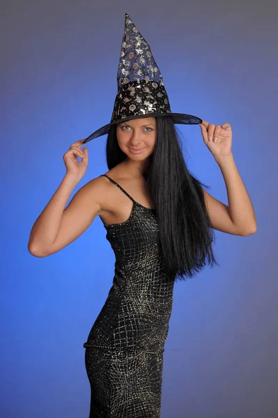 Brunette girl in a black dress and a witch hat on a blue backgro — 스톡 사진