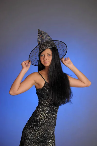 Brunette girl in a black dress and a witch hat on a blue backgro — Stock Photo, Image