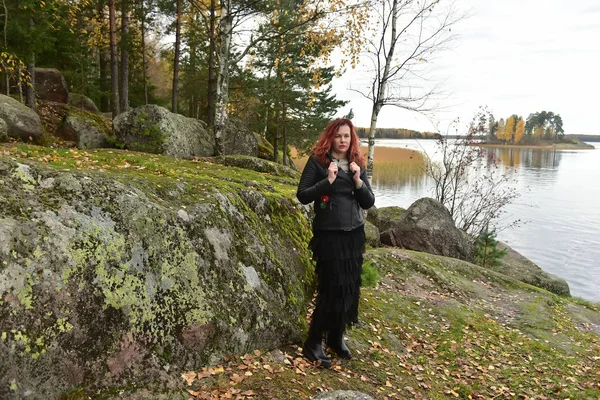 红头发的女人穿着黑色皮夹克 穿着裙子 站在一个石头堆里的牛群公园里 — 图库照片