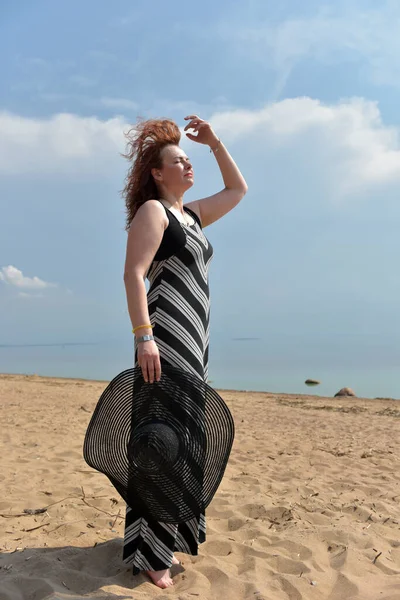穿着条纹连衣裙的女人 手拿着一顶帽子 站在大海的背景上 — 图库照片