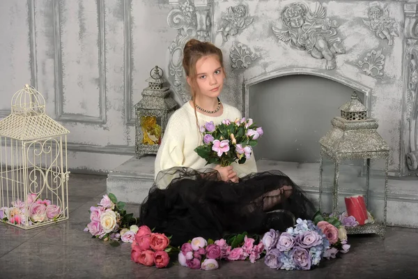 身穿白色毛衣和黑色裙子的漂亮姑娘坐在壁炉边的地板上 鲜花盛开 — 图库照片
