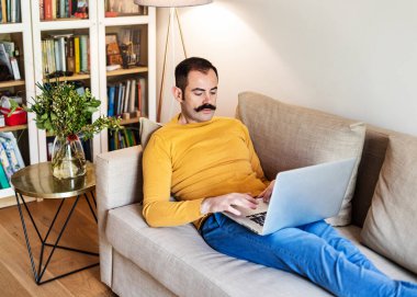 Yetişkin bir adam oturma odasında kanepede oturuyor ve bilgisayarla uğraşıyor.