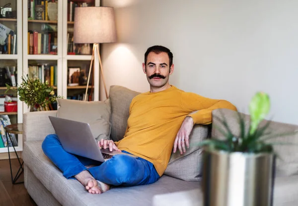 Yetişkin bir adam oturma odasında kanepede oturuyor ve bilgisayarla uğraşıyor.