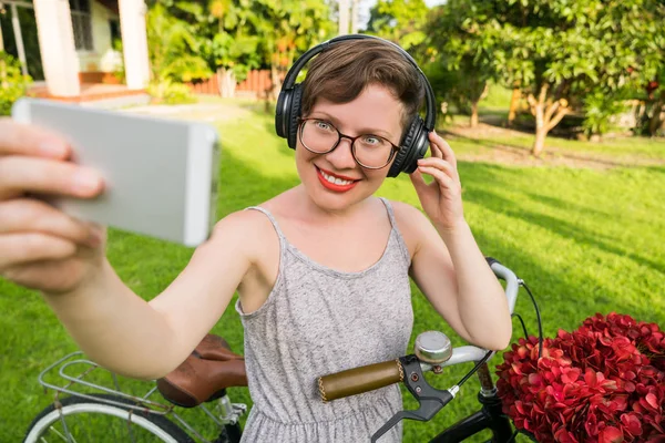 Summer leisure. Pretty young woman in glasses and headphones walking with bicycle. Girl with red lipstick makes selfie photo of yourself for your blog on social networks.
