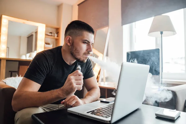 Joven hombre de negocios que trabaja frente a la computadora en casa y fuma un cigarrillo electrónico - freelance y concepto de tecnología . — Foto de Stock