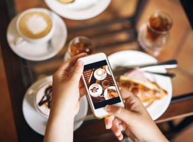 Bir kadın bir telefonu tutuyor. İki Kişilik Kahvaltı: bir kruvasan jambon, kahve, bir serinletici içecek, tatlılık ile.