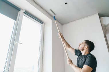 Onun elinde bir silindir bir erkekle tavan penceresi evinin yakınındaki gri renkte boyar. Repainting duvarlar ve tavan oda iç onarmak.
