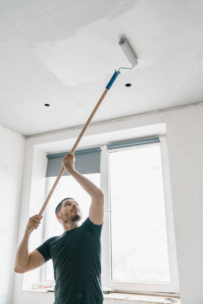 a man with a roller in his hand on a long stick paints the ceiling in gray at the window. Repair in a bright room.