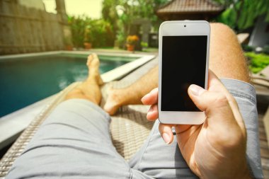 Adam onun smartphone ile boş bir ekran arka plan havuz ve bir şezlong içinde kendi ayakları üzerinde elinde tutuyor. Lüks dinlenme ve teknoloji.