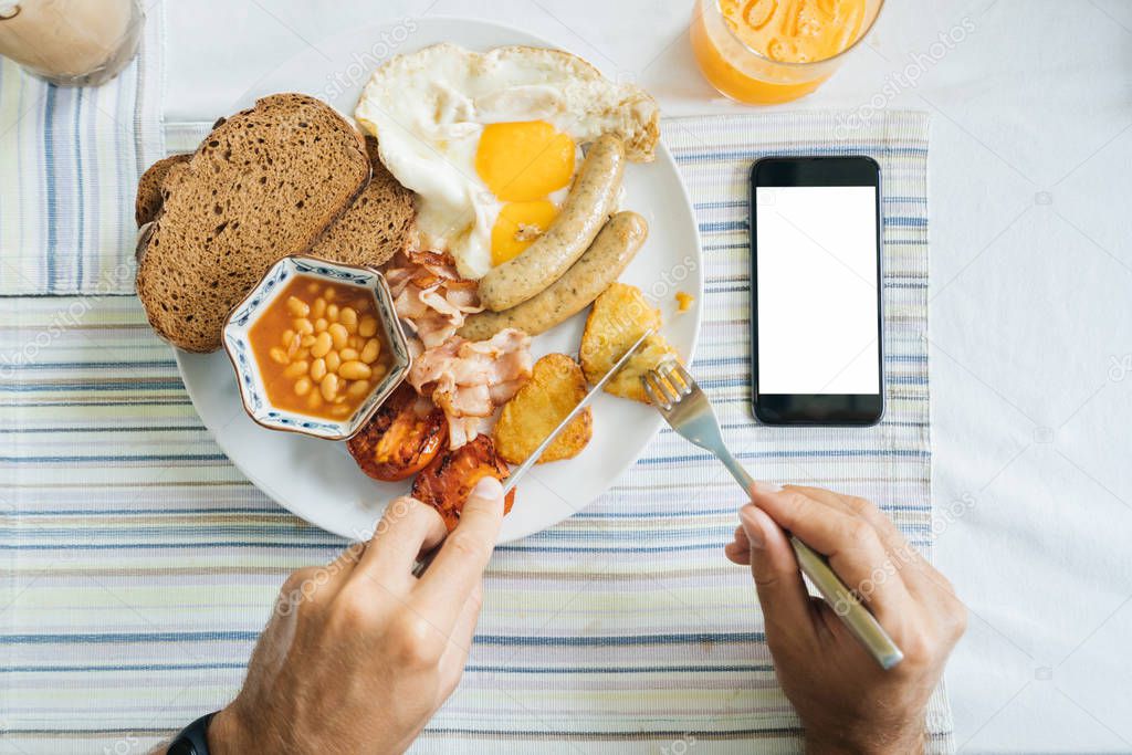 man has breakfast, hands with a knife and a fork, a dish and a smartphone with a blank screen. Calorie 