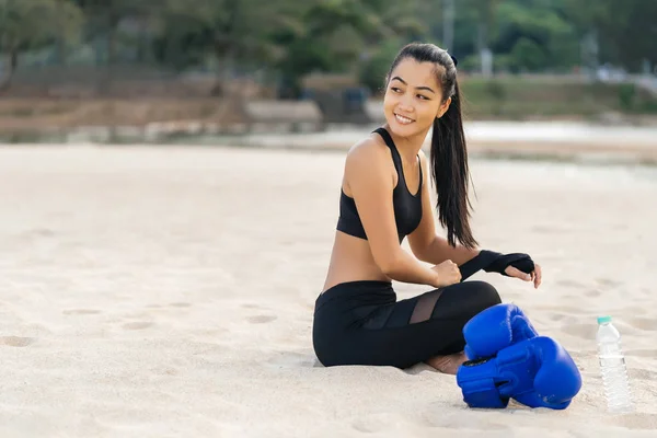 Asian athletic woman a fighter sitting on the beach before boxing training. Healthy lifestyle and summer mood. Closeup portrait with a smile looks away.