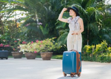 Palmiye yapraklarının arka planında büyük bir bavulu olan şapkalı güzel bir kadın transfer olmayı bekliyor. Tatil ve yaz tarzı.