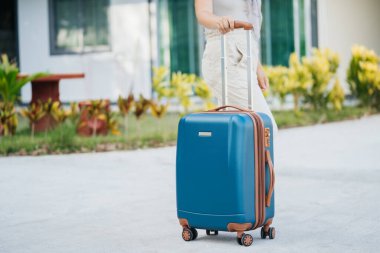 Turist mavisi bavul, bir kadın eli tarafından tropik bir evin arka planında tutulur. Seyahat ve tatil zamanı..
