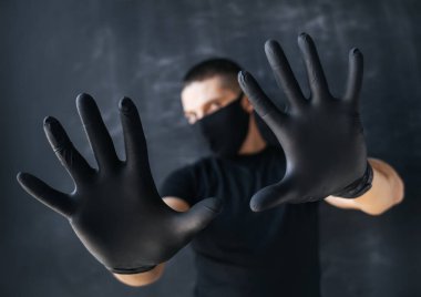 Siyah maskeli ve tıbbi eldivenli bir adam koyu bir arka planda elleri ile durduğunu gösteriyor. Koronavirüs enfeksiyonuna karşı korku ve koruma kavramı. COVID-19