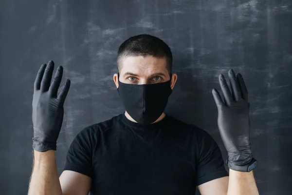 A man in a black mask and medical gloves shows his hands on a dark background. Coronavirus epidemic protection and protection concept.