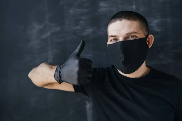 A man in a black mask and medical gloves shows thumb up, on a dark background. The concept of positive protection against coronavirus infection.