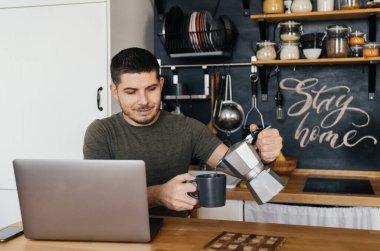 Adam ev mutfağında dizüstü bilgisayarda çalışıyor ve kendine kahve dolduruyor. Uzak çalışma.