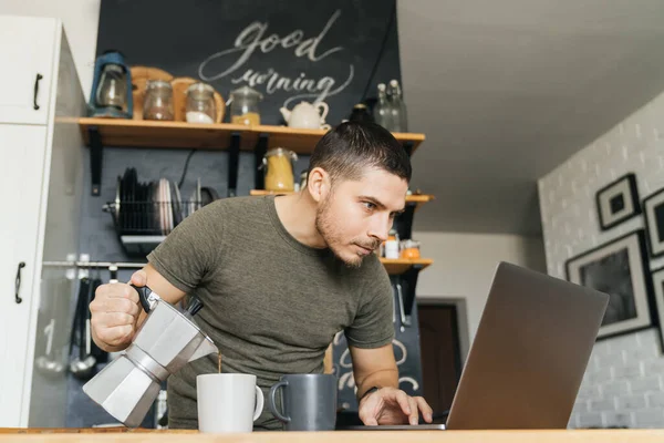 Attractive young man reads news on the Internet and pours himself coffee in the interior of a home kitchen. Morning, distant work.